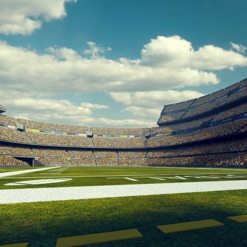 A wide angle panoramic image of a outdoor american football stadium full of spectators under blue sky. The image has depth of field with the focus on the foreground part of the pitch. The view from back line of the field.