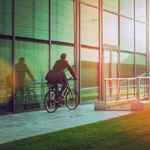 Rear view of a handsome man, on the way to work, riding bicycle beside the modern office building. The man is casually dressed and wears eyeglasses and carries black briefcase hung on shoulder. Blurred motion, copy space has been left.